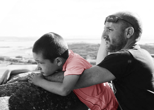 A black and white photo of a boy and a man on a beach lean on a rock together with the boy’s shirt in red. [106]